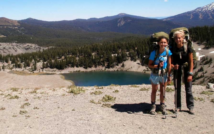 Two students wearing backpacks smile at the camera. There is a blue lake and mountains in the background. 